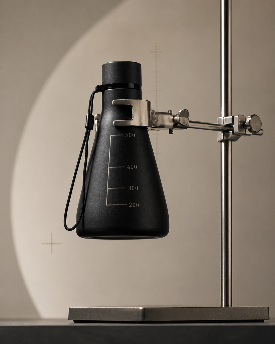 A matte black flask-shaped water bottle held in the clamp of a steel laboratory retort stand, against a beige backdrop lit by a soft arch of light.